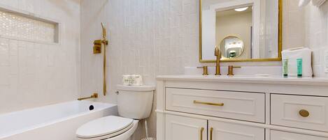 A bathroom with a white bathtub, toilet, and vanity with gold handles.