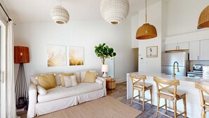 Un espacio habitable moderno con sofá, comedor con sillas, cocina con armarios blancos y lámparas colgantes de mimbre.