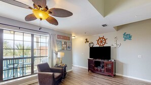 A spacious living room with a large window, a ceiling fan, a TV, and nautical-themed wall decor. - Cozy condo with pools, tennis, gym, mini golf, beach access, & balcony (Panama City Beach)
