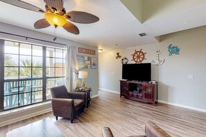 A spacious living room with a large window, a ceiling fan, a TV, and nautical-themed wall decor. - Cozy condo with pools, tennis, gym, mini golf, beach access, & balcony (Panama City Beach)