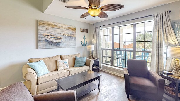 A living room with a beige sofa, a coffee table, a dark armchair, and a ceiling fan. There is a large window with curtains, a painting on the wall, and a small table with a lamp. - Cozy condo with pools, tennis, gym, mini golf, beach access, & balcony (Panama City Beach)