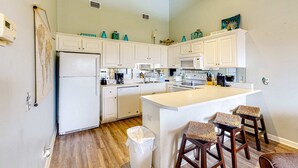 A modern kitchen with white cabinets, a central island with bar stools, a refrigerator, microwave, and a trash can. - Cozy condo with pools, tennis, gym, mini golf, beach access, & balcony (Panama City Beach)