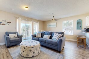 Un salon spacieux avec un canapé gris, un pouf rond orné de motifs marins et un ventilateur de plafond.