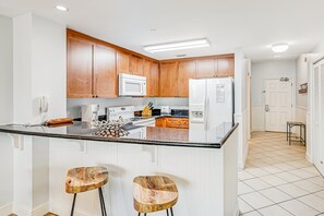 A modern kitchen with wooden cabinets, a central island with stools, and a refrigerator. - Renovated oceanfront condo with pools, hot tub & AC (Panama City Beach)