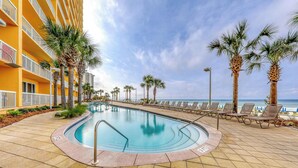 A swimming pool with a curved edge, surrounded by lounge chairs and palm trees, with a building featuring balconies in the background. - Beach-view, high-rise condo with pool - central location (Panama City Beach)