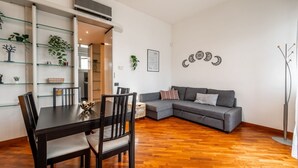 Una sala de estar moderna con piso de madera, un sofá gris, una mesa de comedor con sillas y una estantería de vidrio con plantas.
