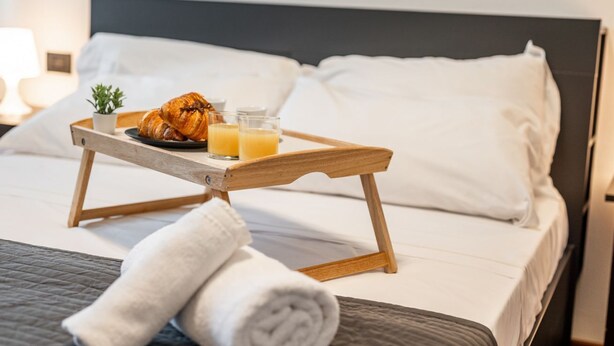 Una cama bien hecha con una bandeja que contiene croissants y jugo de naranja, una toalla enrollada y una pequeña planta en la mesita de noche.