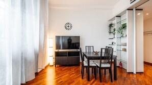 Una sala de estar moderna con piso de madera, una mesa de comedor con sillas, un televisor de pantalla plana y una estantería con plantas.