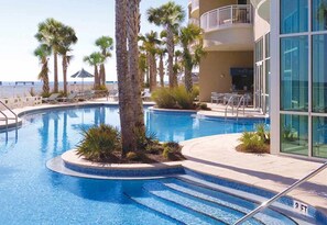 A swimming pool with palm trees, lounge chairs, and a building with large windows. - Stylish oceanfront condo including shared pool and beach access (Panama City Beach)