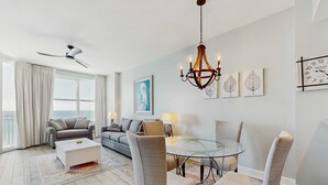 A modern living room with a glass dining table, grey upholstered chairs, a white coffee table, and a ceiling fan. - Stylish oceanfront condo including shared pool and beach access (Panama City Beach)