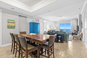 A dining area with a wooden table and chairs, a blue cabinet, and a view of the ocean through a large window. - All-suite condo with Gulf views, beach access, balcony, pool, spa, gym, & AC (Panama City Beach)