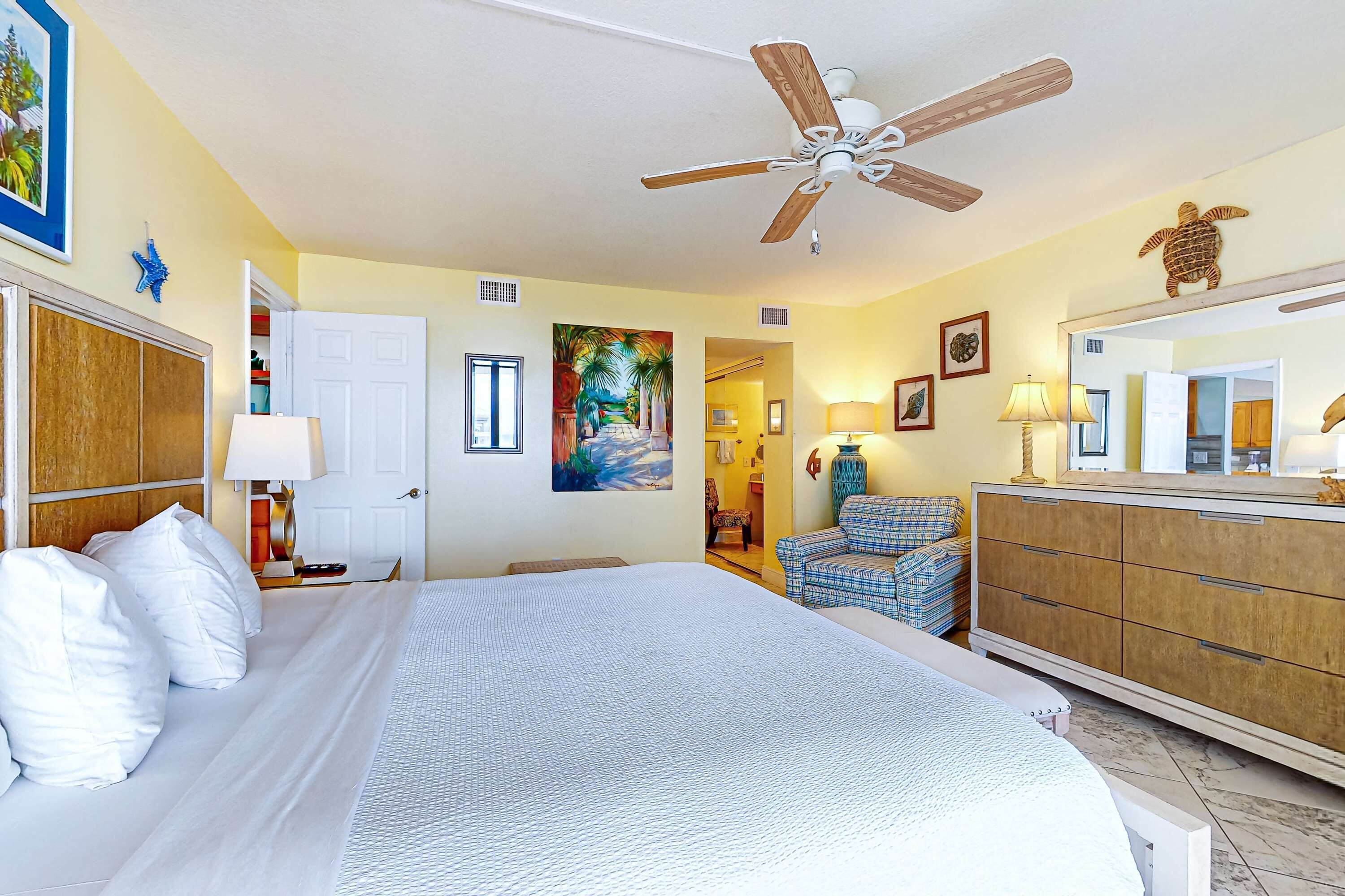 Une chambre d’hôtel avec un grand lit, un ventilateur de plafond, une commode, une chaise et un tableau représentant un paysage tropical.