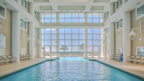 Piscina interna com vista ampla através de grandes janelas, cadeiras de descanso e placa indicando área de mergulho.