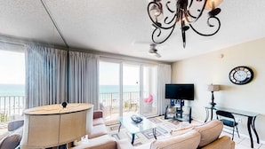 Uma sala de estar ampla com sofá, mesa de centro, TV e uma varanda com vista para o mar.