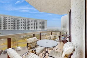 A balcony with a round table and cushioned seating, overlooking a parking area and a large building. - Gulf view resort condo w/ balcony, shared pool/hot tub/tennis & beach access (Miramar Beach)