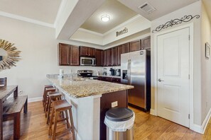 A kitchen with a granite countertop, wooden stools, stainless steel appliances, and a white door. - Condo in Miramar Beach with a pool & stellar views (Miramar Beach)