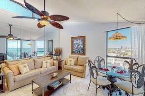 A modern living room with a sofa, coffee table, and dining table with chairs. There is a ceiling fan, large windows with a city view, and a painting on the wall. - 8th Floor Comfortable gulf view Condo, beach setup included, Near dining (Miramar Beach)