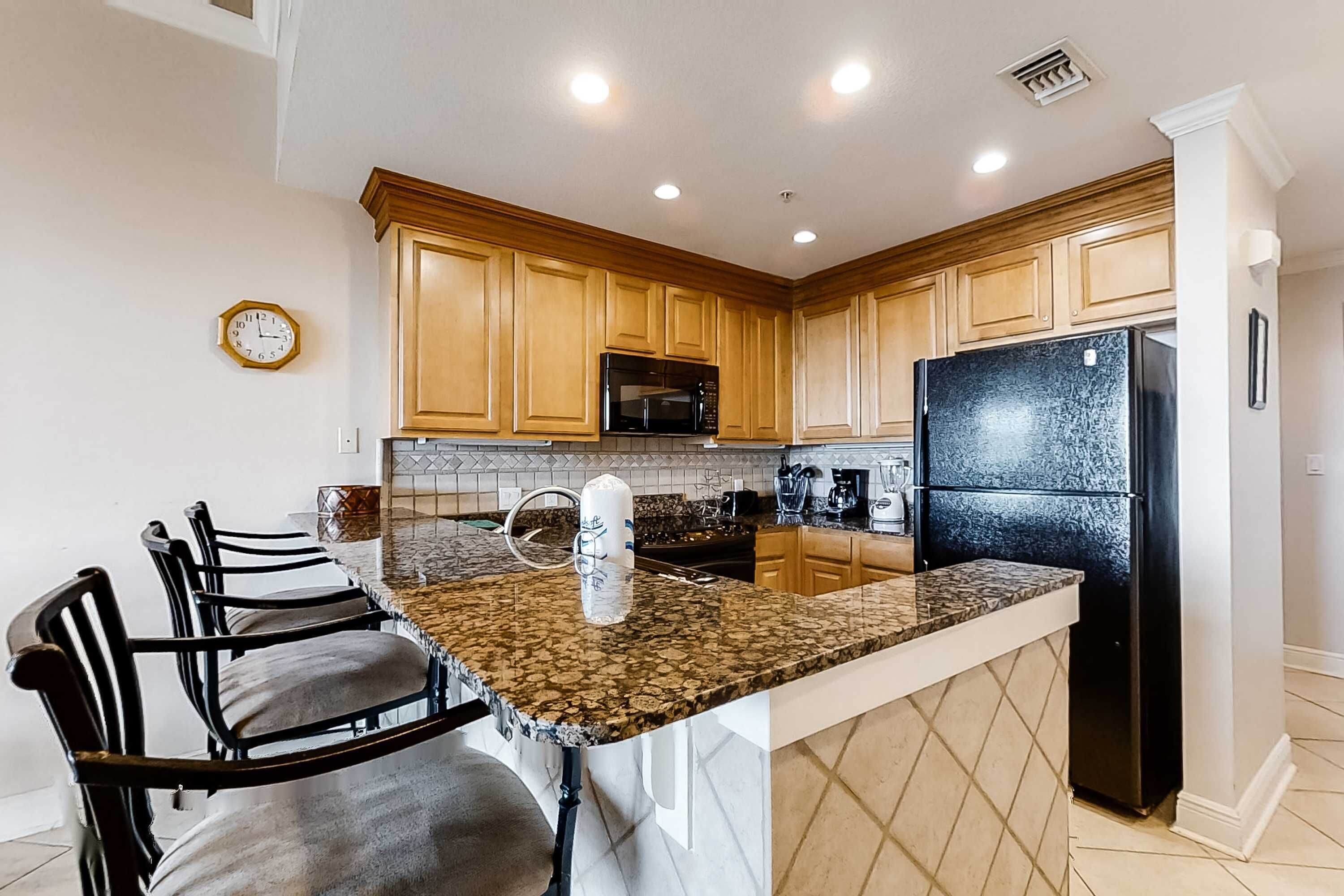 A kitchen with wooden cabinets, a granite countertop, a black refrigerator, and a clock on the wall.