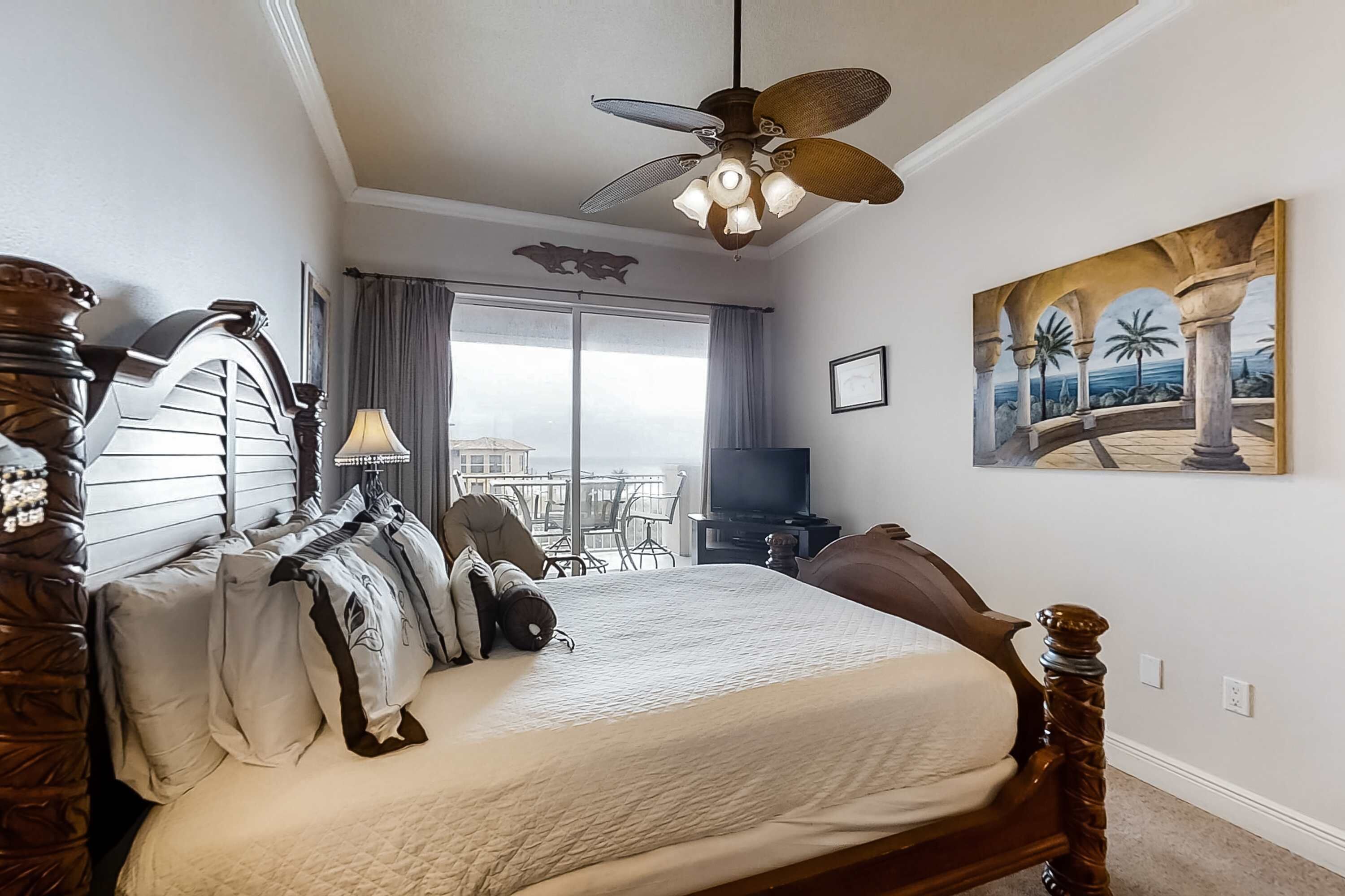A bedroom with a large bed, a ceiling fan, a painting, and a view of the outdoors.