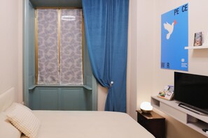 Une chambre à coucher avec un lit, une télévision, une petite table avec une lampe, une fenêtre avec des rideaux et un mur orné d’une affiche sur le thème de la paix.