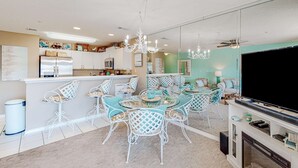 A modern kitchen with white cabinets, a refrigerator, and a dining area with a glass table and white chairs. - Pet-friendly, pools, hot tub & balcony with Gulf-side calm (Destin)