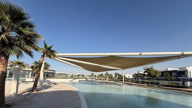 A swimming pool with a shaded area, palm trees, and modern buildings in the background. - Elegant Villa with Direct Pool/Lagoon Access (Dubai)