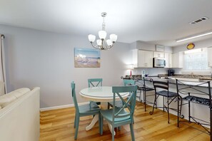 Una cocina moderna con armarios blancos, microondas y un lavamanos. Hay una zona de comedor con una mesa blanca y sillas color turquesa.