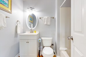 Un baño con lavamanos blanco, espejo e inodoro. Hay un perchero en la pared y una puerta a la derecha.