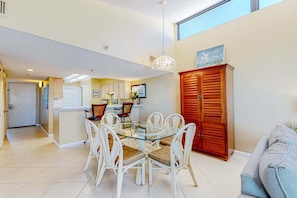 A dining area with a glass-top table surrounded by white chairs, a wooden cabinet, and a grey sofa. - 8th Floor Open, Updated, condo, Beach service included, Near Everything (Miramar Beach)
