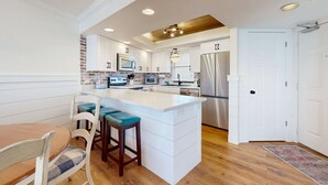 A modern kitchen with white cabinetry, a central island with bar stools, a brick accent wall, and a wooden ceiling. - Gulf-front condo with balcony, ocean view, W&D, pool, hot tub & tennis (Miramar Beach)
