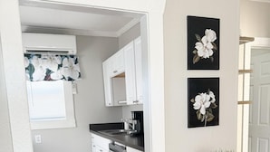 Cocina compacta con armarios blancos, cafetera y mesa de comedor con sillas. Hay dos cuadros de flores en la pared y una planta en maceta sobre la mesa.