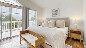 Quarto com uma cama grande, um banquinho, uma mesinha de cabeceira, janela com cortinas e vista para uma varanda.