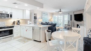 Una cocina moderna con armarios blancos, microondas integrado y cafetera. Cuenta con una zona de comedor con una mesa redonda y sillas blancas.