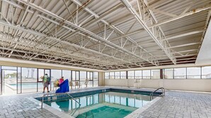 An indoor swimming pool with a tiled floor, a metal roof structure, and large windows offering views of the outside.