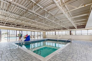 An indoor swimming pool with a tiled floor, a metal roof structure, and large windows offering views of the outside.