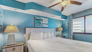 A bedroom with a large bed, a ceiling fan, and a view of the beach through a window.
