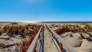 Ein hölzerner Steg führt zu einem Strand mit Dünen und spärlicher Vegetation.