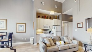 A modern living room with a grey sofa, a dining table with chairs, and a kitchen area in the background. - Spacious condo with large living area, private balcony & shared pool (Santa Rosa Beach)