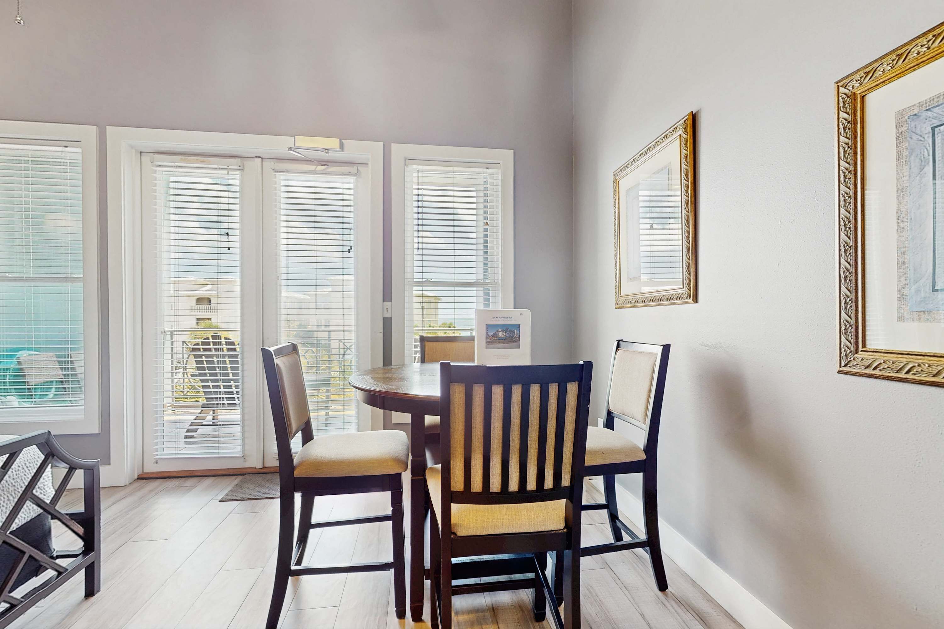 A dining area with a round table, four chairs, and a view through large windows with blinds.