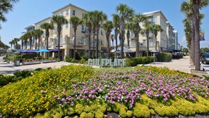 A hotel with a central courtyard featuring a flower garden with pink and yellow flowers, surrounded by palm trees and a building with balconies. - Spacious condo with large living area, private balcony & shared pool (Santa Rosa Beach)