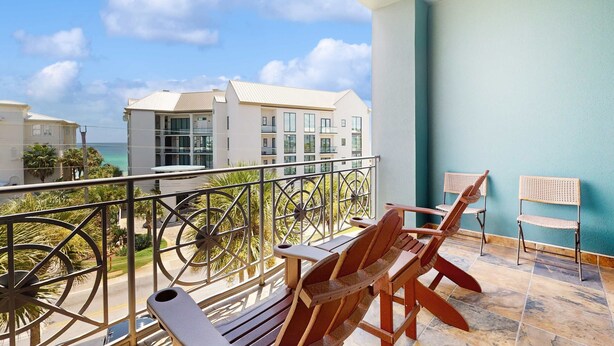 A balcony with wooden chairs and a table, overlooking a building and a view of the sea. - Spacious condo with large living area, private balcony & shared pool (Santa Rosa Beach)
