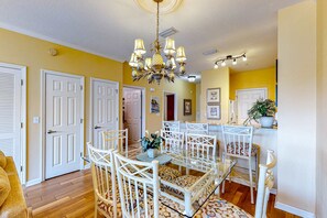 A dining area with a glass table and white chairs, a chandelier, and a kitchen area with a plant. - Dog-friendly condo with pool, hot tub, private balcony, AC, & washer&dryer (Miramar Beach)