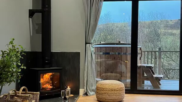 Ein gemütliches Wohnzimmer mit Kaminofen, einem großen Fenster mit Blick auf eine Terrasse und Bäume sowie einer komfortablen Sitzecke.
