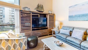 A modern living room with a large TV, a sofa, a coffee table, and a view of the sea through the window. - Spacious, beachfront home w/ shared outdoor pool, hot tub, & beach access (Miramar Beach)