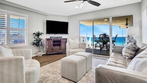 A spacious living room with a ceiling fan, a flat-screen TV, a fireplace, and a balcony with a view of the ocean. - Big 'O Beach Miramar/Destin (Miramar Beach)