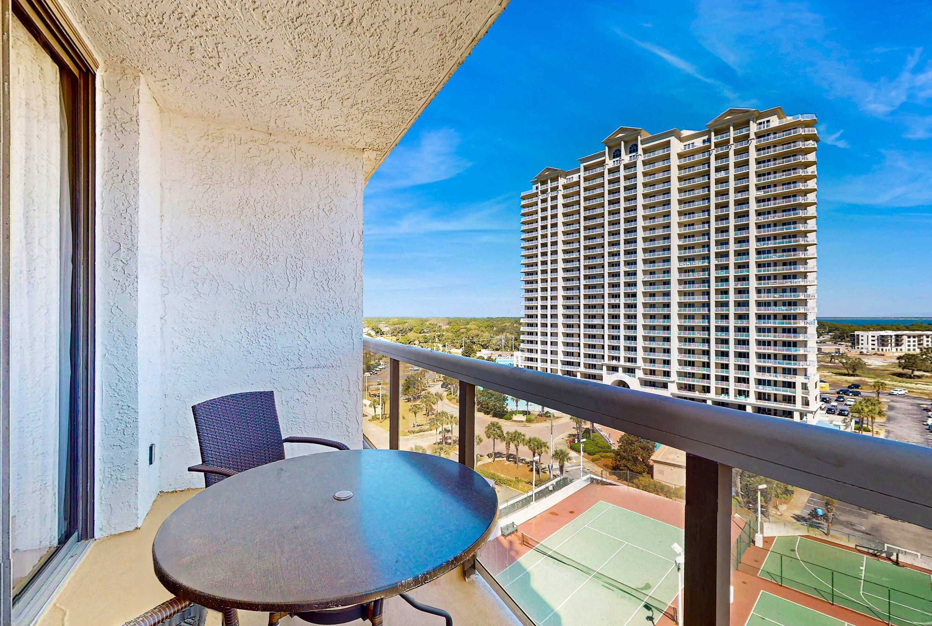 A balcony with a table and chair, overlooking a tennis court and a high-rise building.