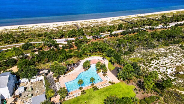 An aerial view of a resort with a swimming pool surrounded by greenery, a beach, and a road.