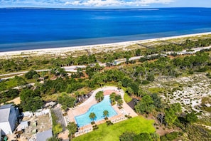 An aerial view of a resort with a swimming pool surrounded by greenery, a beach, and a road.