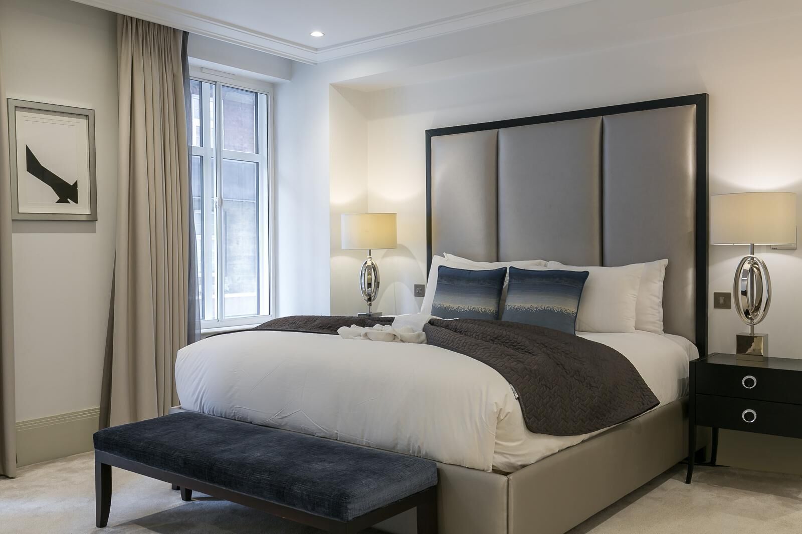 A modern bedroom with a large bed, a bedside table, a bench, and a window with curtains.