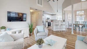 Un salón amplio con ventilador de techo, televisor montado en la pared, comedor con mesa y sillas, y una cocina al fondo.